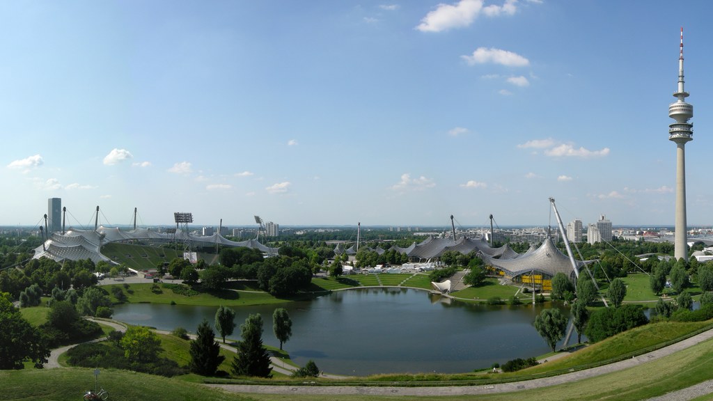 Aufnahme vom Olympiapark München zeigt asphaltierte Laufwege, einen Betontunnel unter der Spirale und die filigrane Dachkonstruktion aus Stahl und Glas im Hintergrund.