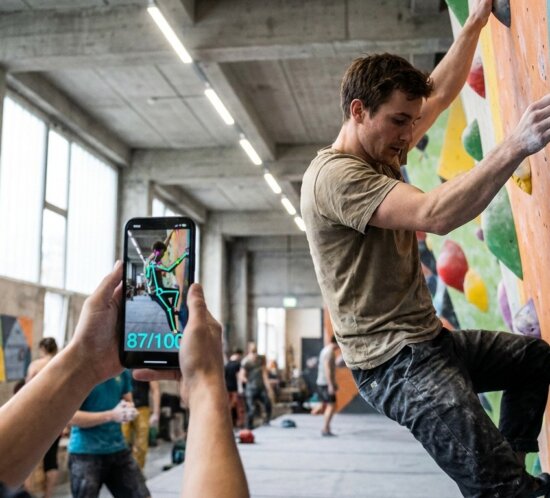 Eine Person filmt einen Boulderer an der Kletterwand, auf dem Smartphone ist eine Pose-Analyse zu sehen.