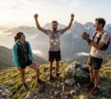 Drei Trailrunner stehen bei Sonnenaufgang auf einem Berggrat, einer hebt jubelnd die Arme.