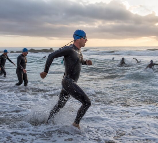 Triathleten in Neoprenanzügen und blauen Badekappen laufen bei Wellengang ins Meer.
