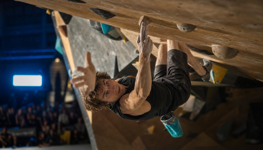 Kletterer hängt in einer überhängenden Boulderwand und greift nach einem Griff vor Publikum.