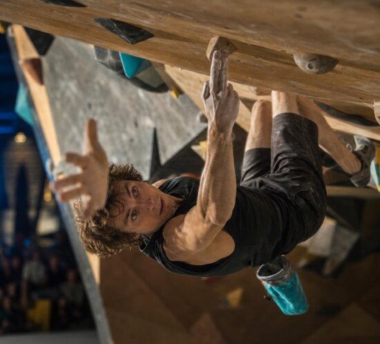 Kletterer hängt in einer überhängenden Boulderwand und greift nach einem Griff vor Publikum.