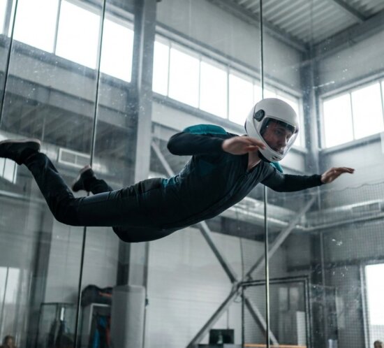 Indoor-Skydiver mit Helm schwebt horizontal in einem gläsernen Windkanal einer Trainingshalle.