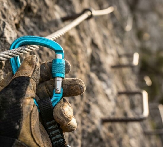 Hand mit Kletterhandschuh befestigt einen blauen Karabiner an einem Stahlseil am Fels.