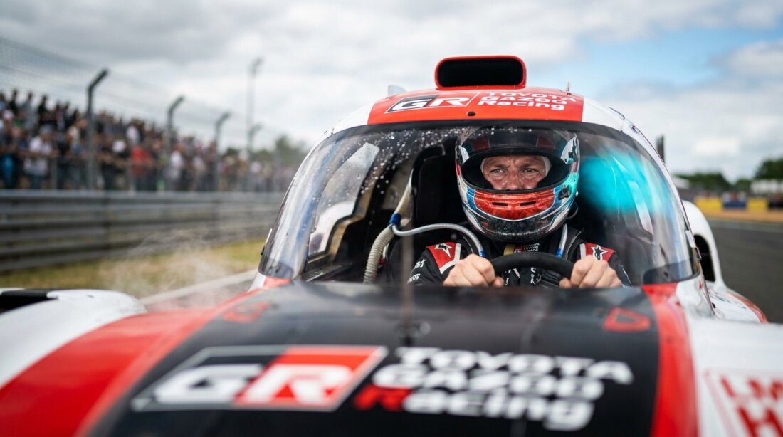 Rennfahrer mit Helm sitzt im Toyota-Gazoo-Racing-Wagen auf der Strecke, Zuschauer im Hintergrund