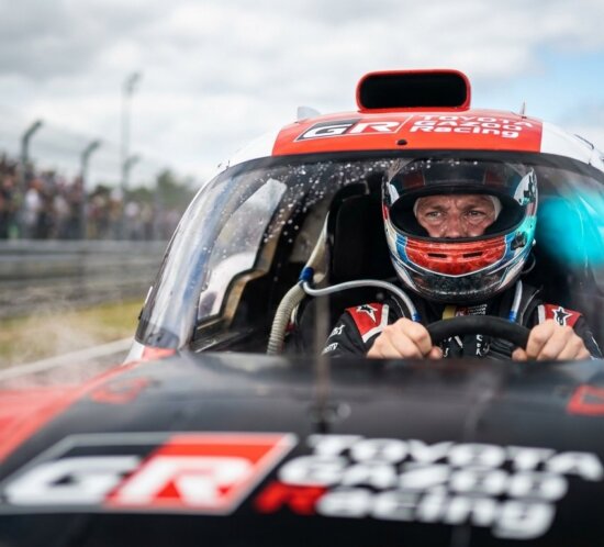 Rennfahrer mit Helm sitzt im Toyota-Gazoo-Racing-Wagen auf der Strecke, Zuschauer im Hintergrund