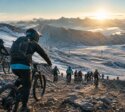 Mountainbiker starten auf einem felsigen Schneehang vor Bergpanorama im Sonnenaufgang.