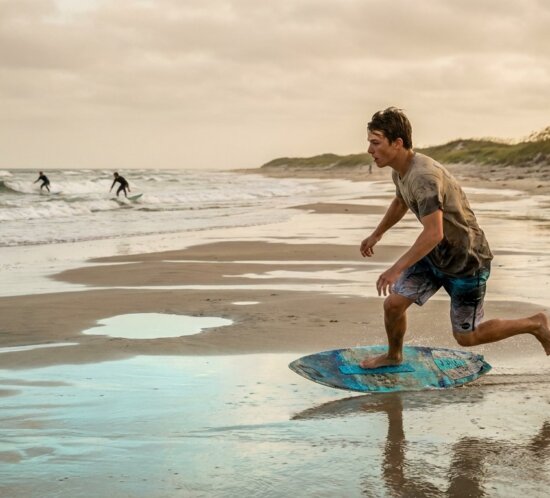 Junger Skimboarder läuft am nassen Strand auf seinem Board, während im Hintergrund Surfer im Meer sind.
