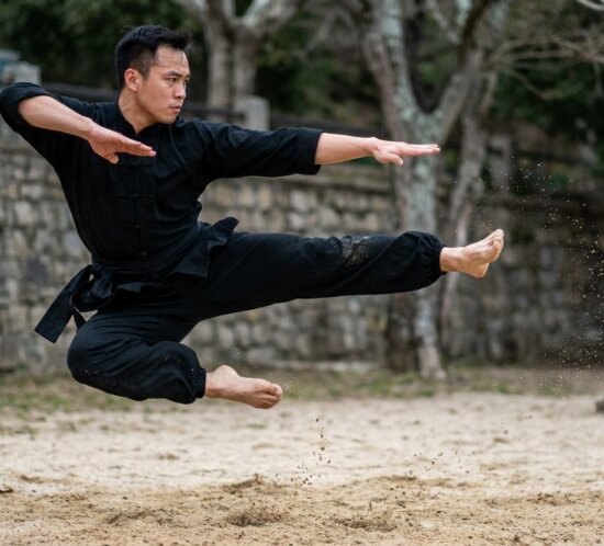 Wushu-Sportler in schwarzer Kleidung springt mit ausgestrecktem Bein über sandigem Boden.