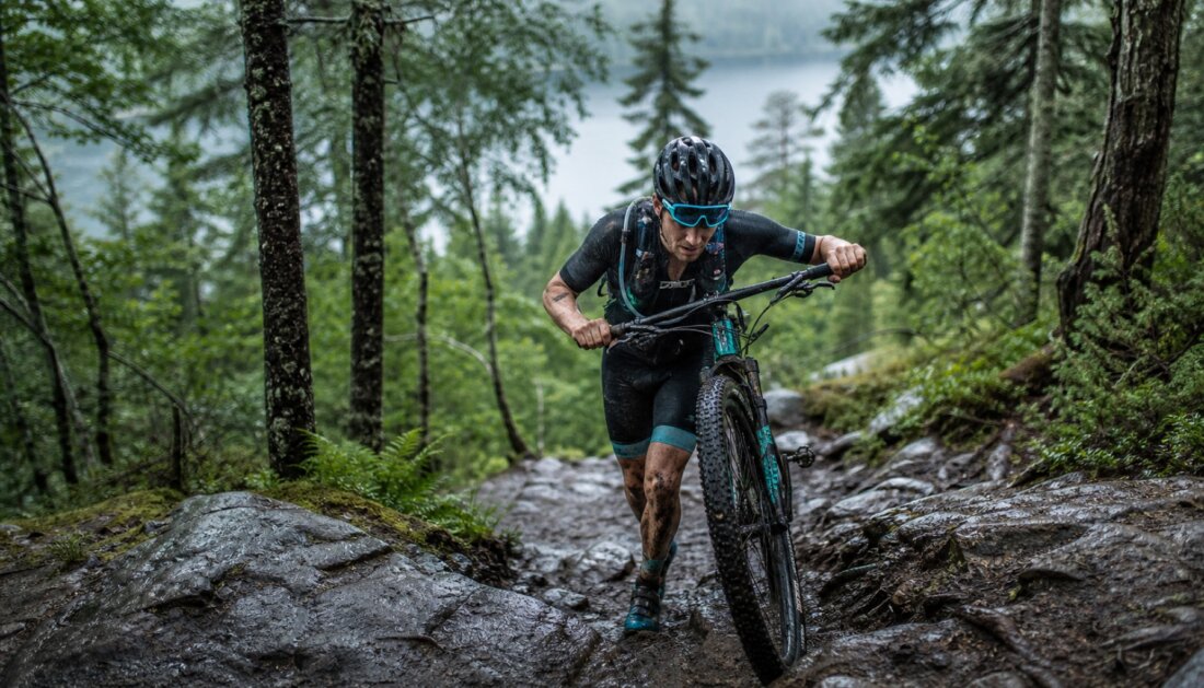 Ein Mountainbiker schiebt sein Rad auf nassem, steinigem Waldtrail zwischen Bäumen, hinten ein See