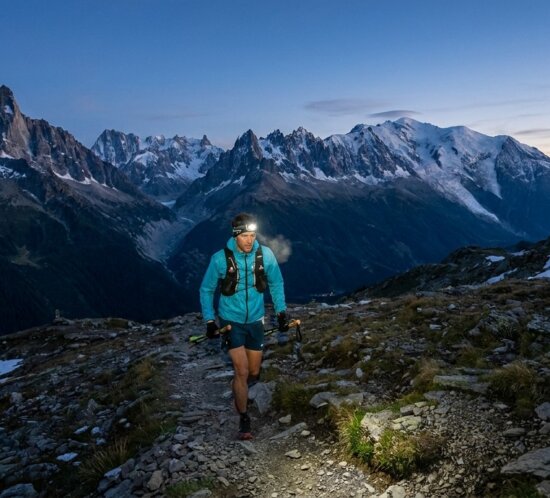 Trailrunner mit Stirnlampe steigt bei Dämmerung über einen steinigen Alpenpfad vor schneebedeckten Gipfeln