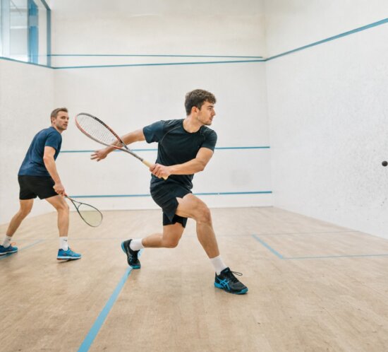 Zwei Männer spielen Squash auf einem hellen Court, einer schlägt den schwarzen Ball.