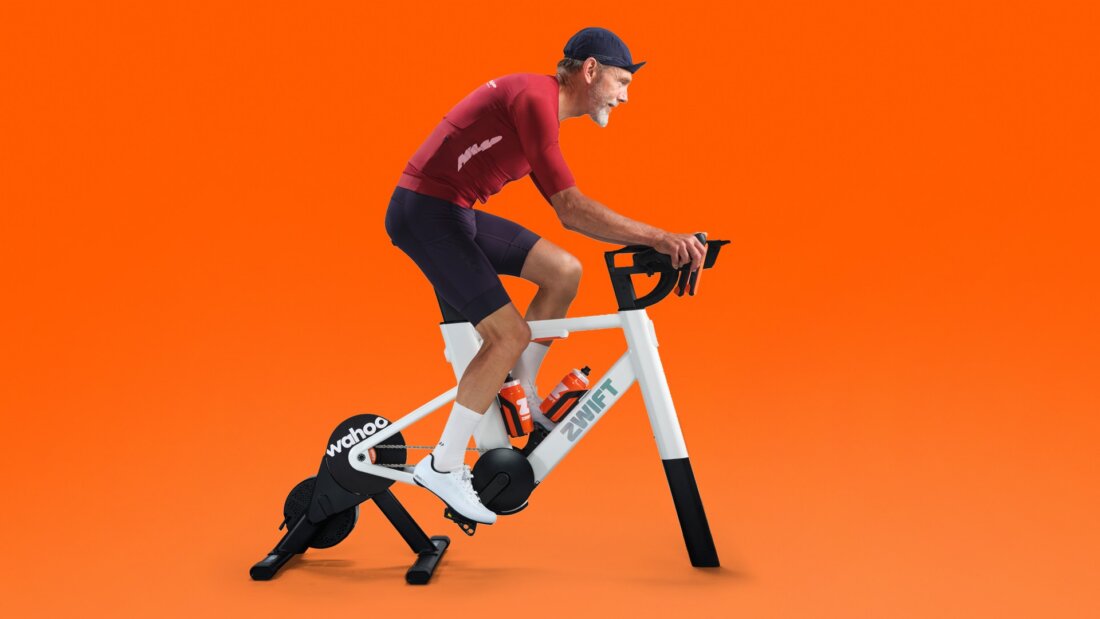 Fahrer auf dem Zwift Ride V2 mit Wahoo Kickr Core vor oranger Fläche (Pressebild Zwift)